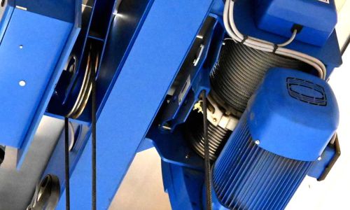 Nahaufnahme eines blauen Seilzugs mit Elektromotor und Umlenkrollen, montiert an einem Hallenkran.